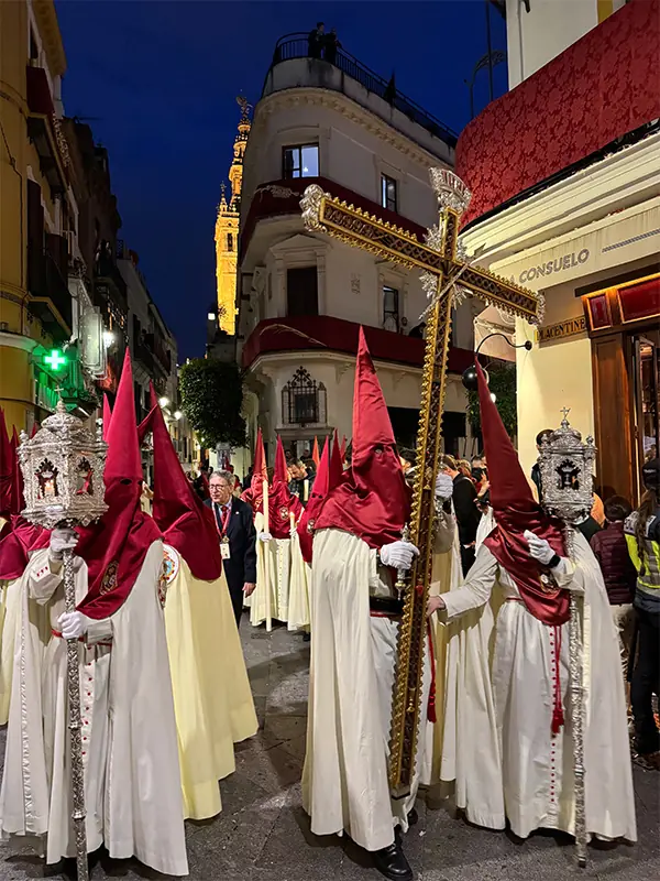 Nazarenos w czerwonych szatach niosący złoty krzyż podczas nocnej procesji Wielkiego Tygodnia w Hiszpanii.