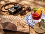 Wakacyjny harmonogram kursów hiszpańskiego i portugalskiego online. Na drewnianym stole leży mosiężny zegar słoneczny, hiszpański wachlarz, aparat fotograficzny oraz napój na portugalskiej płytce azulejo.